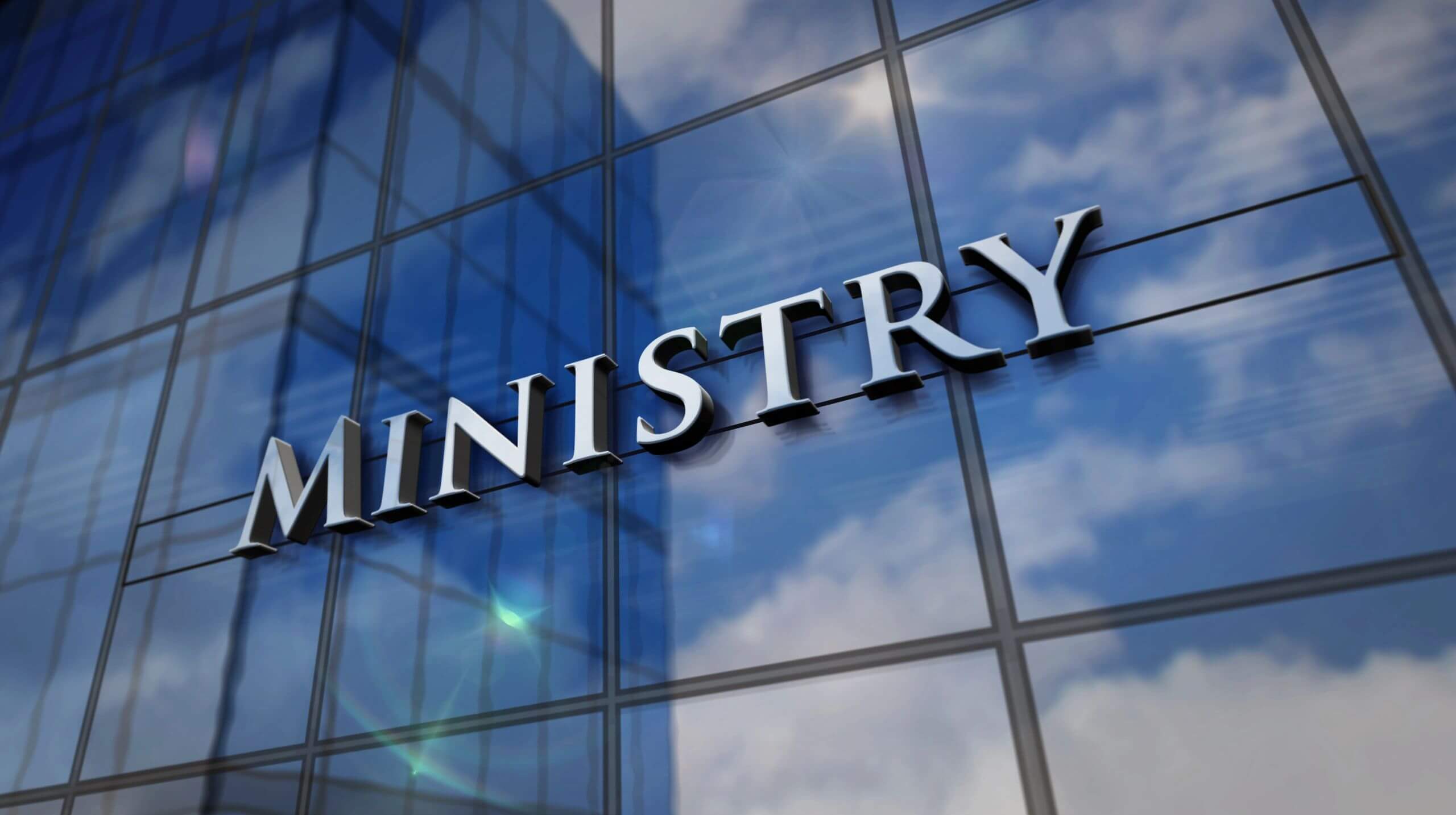 Ministry sign reflected on a glass building under a blue sky.