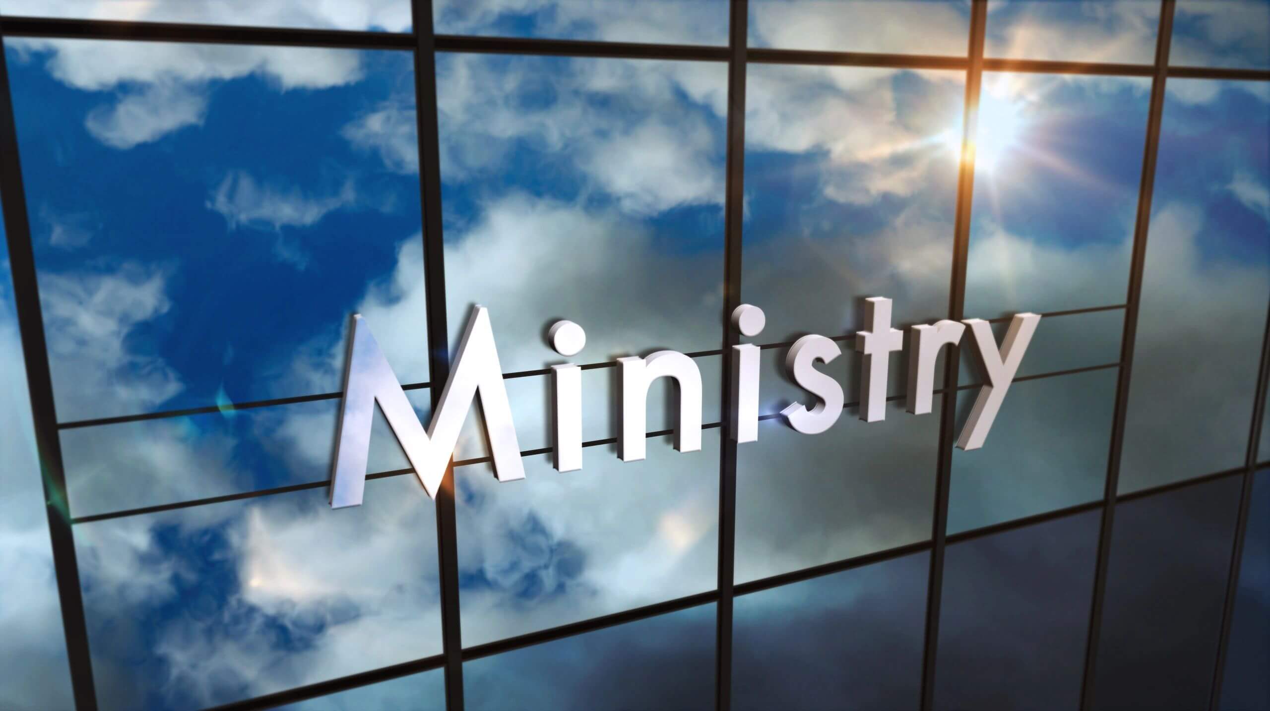 Reflective glass panel with the word 'Ministry' against a blue sky with clouds.