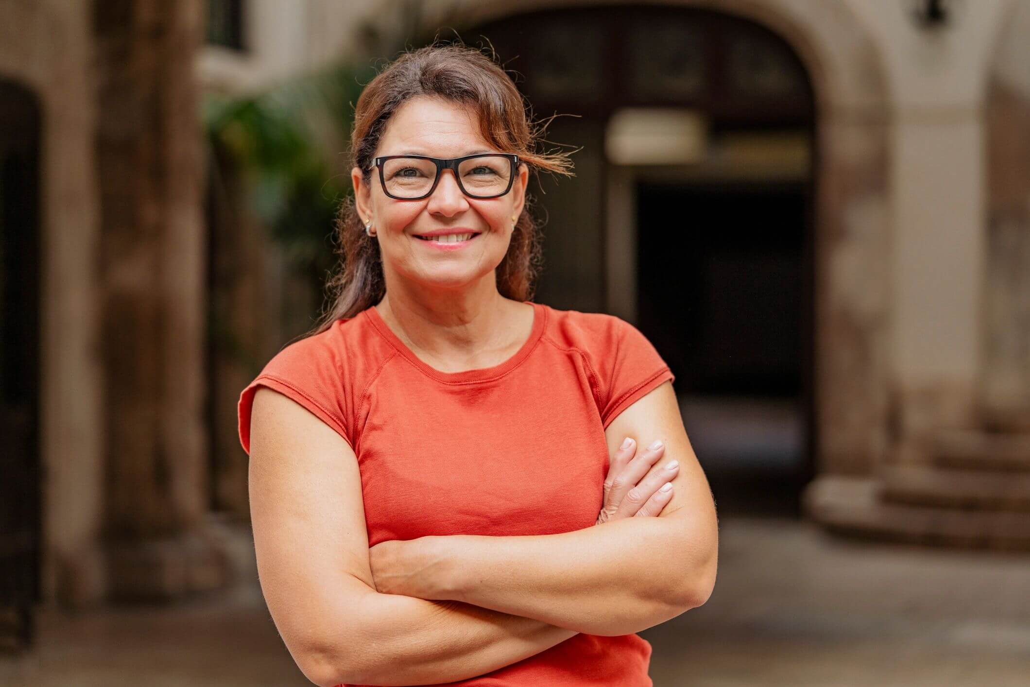 Smiling woman with glasses and crossed arms in an outdoor setting.
